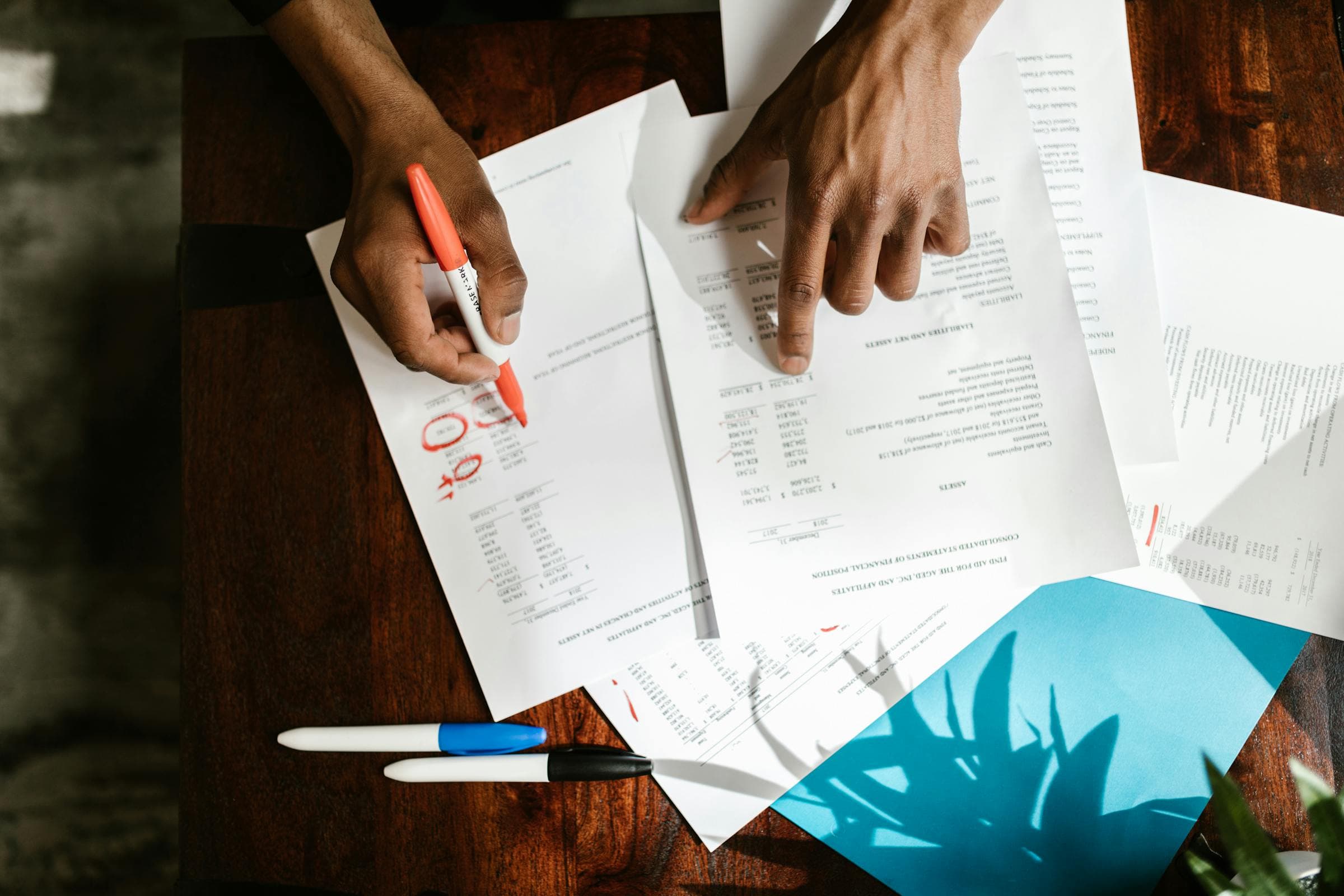 Overhead view of hands marking up financial statements and reports on a wooden table with colored markers